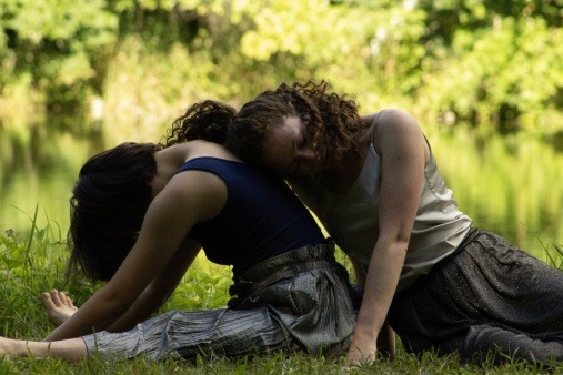 two dancers sit in the grass. One dancer leans over her legs, while the other dancer rests on the first dancer's back.