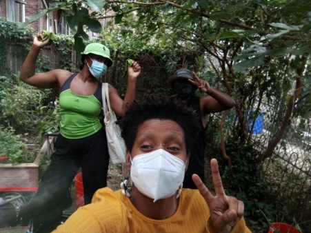 3 Black people in masks, throwing up a hand and smiling in a garden