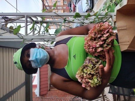 a Black woman in a light blue mask, green bucket hat, & green tank top, with hydrangea flowers in her hands, standing on a porch 