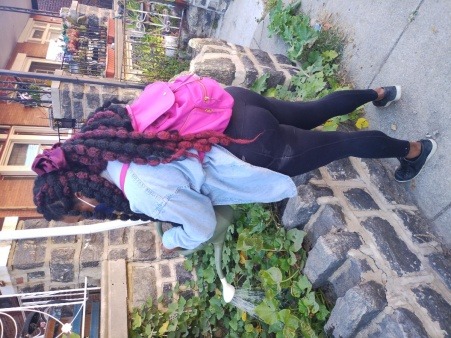 the view from behind of a Black person with long pink and black hair, a jean shirt, leggings, & pink backpack watering a raised stone bed from a pail