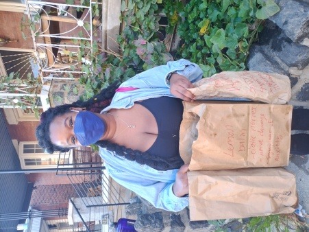 a Black person with long pink and black hair, a jean shirt, leggings, & a blue mask holding 3 paper bags in their hands 