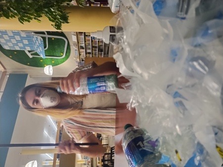 a white woman with glasses, a white mask, & a striped shirt, holding a metal rod to ecobrick a plastic bottle at a table covered in plastic pieces, at a library 