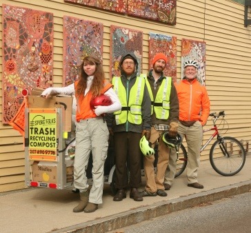 4 people stand in front of a bike attached to a trailer. The trailer is loaded with trash bags and has a sign that reads Spoke Folks