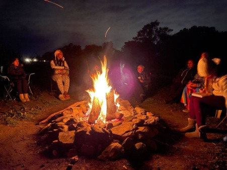 Glow of a fire at night illuminates those sitting around it with the sky lit in the background