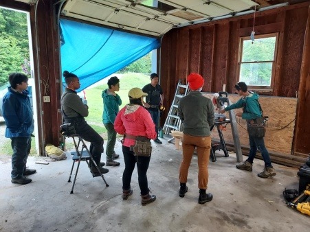 A group of people in an open garage, all focusing on a teacher explaining the use of a tool.