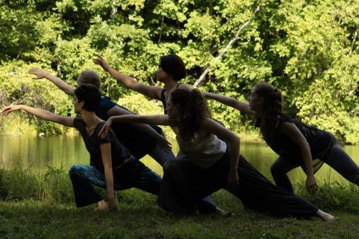 Five dancers outdoors lean to the right in a lunge, with their arms outstretched. 