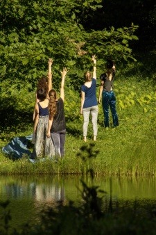 Five dancers outdoors stand backwards, in front of a pond, with their right arms outstretched above their heads.