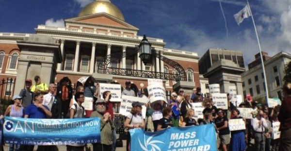 Alternate Text: A crowd of protesters, including the Climate Action Network that LexCAN is a part of, stands outside the statehouse, holding signs supporting climate action in Massachusetts.