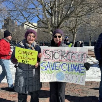 Two LexCAN Board Members, Fran Ludwig and Ricki Pappo, protest in Boston in favor of climate action. Fran Ludwig holds a yellow sign that says, 'Change the Climate in DC', and Ricki Pappo holds a sign that says, 'Stop the Coup, Save the Climate'