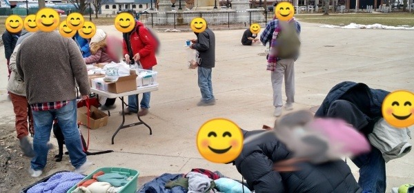 About 20 people with faces covered by smiley emojis gather around folding tables piled with items such as clothing and food. 