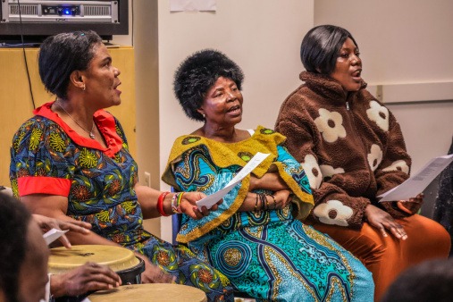 Three members of the Congolese Choir at a 2026 All the Rivers rehearsal Three members of the Congolese Choir are shown seated, wearing colorful traditional African clothing and singing during an All the Rivers Rehearsal