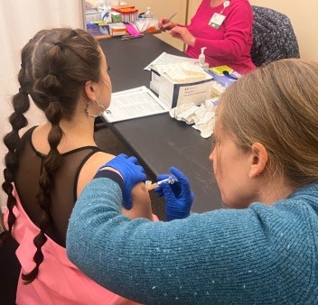 a member with bubble braids and a pink jacket is getting a vaccine from a nurse in a blue sweater next to a table with supplies on it