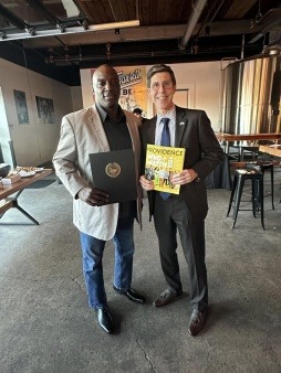 Arthur Johnson wearing a tan blazer, black shirt and jeans standing next to Providence Mayor Brett Smiley who is wearing, glasses, a grey suit. Arthur is holding a City Citation and Mayor Smiley is holding a copy of Providence Monthly Magazine