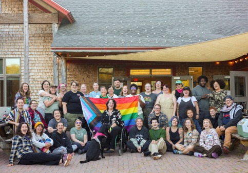 A group of 30-sum campers sit and stand together in front of a large shingled building. In the center of the group, several campers hold up a rainbow Pride flag.
