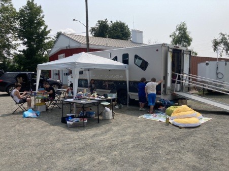 The joy truck is parked in a dirt parking lot with a pop up tent along the side, cushions on the floor next to th ramp. Young children stand at the chalkboard alongside the struck and sit undr the tent making art and playing games. 