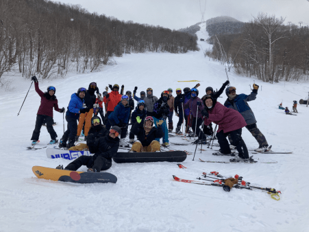 A group of 23 people in ski and snowboard gear are posing together, some sitting in the front, others standing around them, some with their hands up, at a midmountain on snow location, with an open snow trail and top of the tram visible behind them.