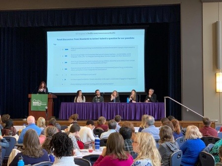 a presentation taking place in a large room with one person at a podium and several seated panelists at a table in the background with a large seated audience in the foreground