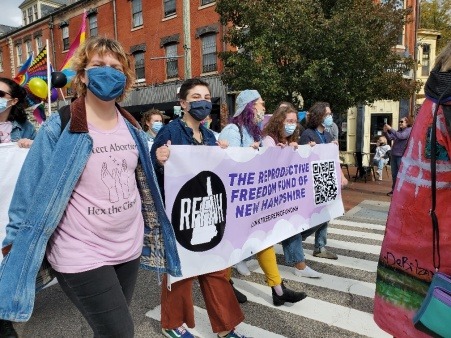 Group of people who appear to be marching in a parade, holding a banner that says "Reproductive Freedom Fund of New Hampshire". Person on the left is wearing a shirt that says "Protect Abortion, Hex the Cis-tem"