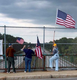 Three New Hampshire Bridge Brigade for Democracy volunteers stand on an overpass holding American flags, facing the highway below. They are participating in a peaceful, community-led visibility action designed to promote civic engagement and public safety.