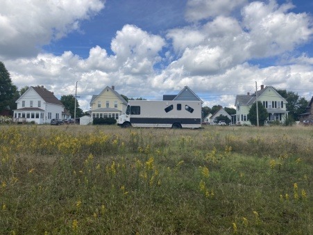 The Joy Truck is parked in the middle of a grassy field in front of a row of houses along a railroad in a town called Derby. It is summer and the clouds are large and puffy and the dandelions are in bloom. 