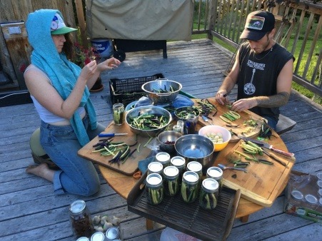 Two white folx, one AFAB, one AMAB, sit around a low table outside on a porch.  The table is covered with multicolored beans, cutting boards, jars being filled and jars already full.  They are making dilly beans.  