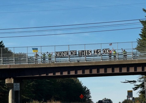 Volunteers stand on a New Hampshire highway overpass holding a large banner reading “NONE OF THIS IS OK,” along with flags and safety gear, during a peaceful NH Bridge Brigade for Democracy visibility action.