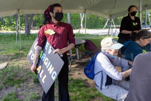 A member of Housing and Climate Justice for Acton holds a sign that says "EV Charging for All" at a picnic outside Acton's annual Town Meeting in May 2022.