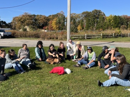 A group of farmers sitting outdoors in a circle point to a CPR dummy wearing a Young Farmer t-shirt