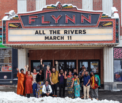 Members of the All the Rivers ensemble gathered under the Flynn marquis announcing the benefit concert Members of All the Rivers smiling and waving under the Flynn marquis listing "All The Rivers March 11"