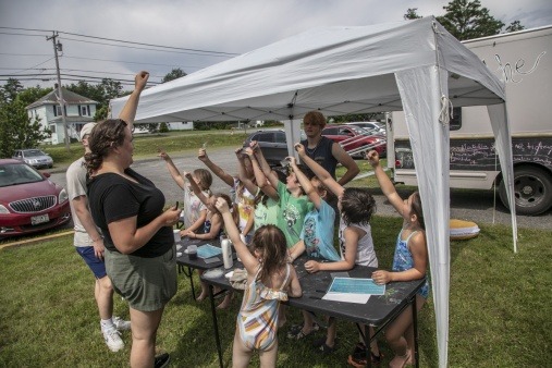 Liza White, graduate student in the Bioengineering department at University of Maine teaches young children about light refraction. They stand under the pop-up tent to the side of the Joy Truck outstretched arms holding small rainbow squares up to the sky. 