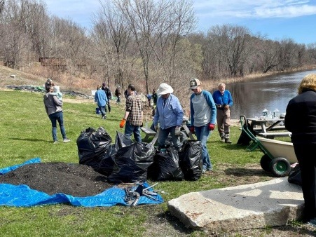 People are standing  on a green lawn along a river bank. The eaves are just emerging on the trees along the riverbank. There are five full contractor trash bags, a wheelbarrow, and a tarp holding wood chips on the lawn