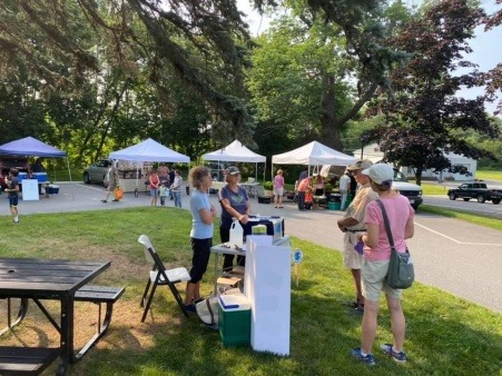 Two adults are standing behind a table are speaking to two other adults. They are located on a lawn at a Farmers Market. There are tents in the background sheltering market vendors .