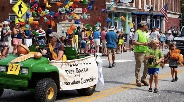 A person drives a green offroad labeled with a banner reading "Hallowell Tree Board and Conservation Commission" in a parade. The vehicle is followed by an adult and a youth handing out tree seedlings to spectators on the parade route.