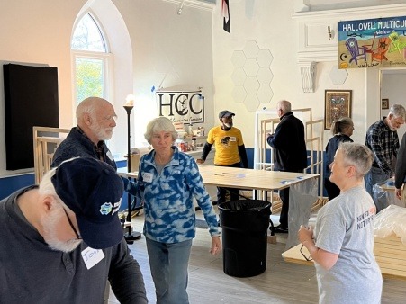 People are speaking to one another and working at tables in a large room. A banner printed with "HCC-Hallowell Conservation Commission" hands on one wall, and a banner printed with "Hallowell Multicultural Center" hands above a doorway. Window inserts at varying stages of completion are stacked at work stations 