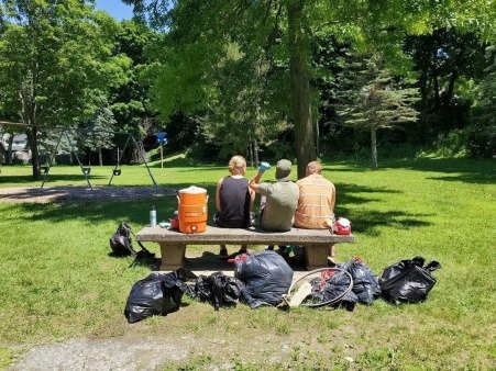 Three Needlepoint organizers sitting on a bench next to a water container in Second Street Park, Bangor. Behind them are seven bags full of items collected from the cleanup which took place in July of 2020. 