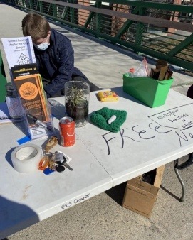 Close up photo of table with "Free Narcan and Needlepoint Sanctuary Of Maine"" written across the top in Pickering Square, Bangor. Placed on top of the table are Needlepoint informational pamphlets, a Bangor-area general resources map, a yellow plastic container filled with instramuscular narcan. An organizer wearing a dark blue coat and light blue mask is seated behind the table.