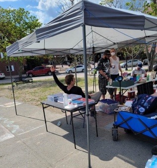 Organizers and friends, some sitting and some standing, under a white canopy and next to two black tables. On top of the table to the right is five boxes of pizza. The other table holds beverages, a box with pipes and straps, containers with mutual aid supplies, and Needlepoint informational pamphlets. A red sharps container sits below the table, next to a black sleeping bag.