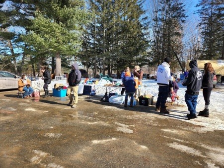 A group of ten people next to two white tables lined with hot beverages and food, mutual aid and harm reduction supplies at Camp Hope in Bangor, Maine. Next to the tables are totes filled with winter clothing. There is some snow in the background.