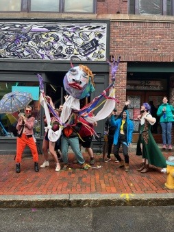 A crowd of people dance around a large puppet, one person holds the head, two hold the hands