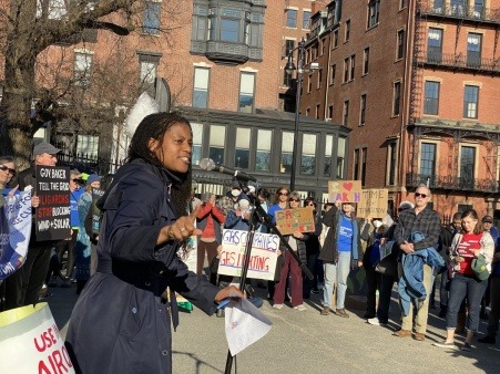 State Senator Lydia Edwards addressing the crowd at a Fix the Grid rally in Boston in March of 2022. Folks around her are holding signs that call for a clean energy transition.