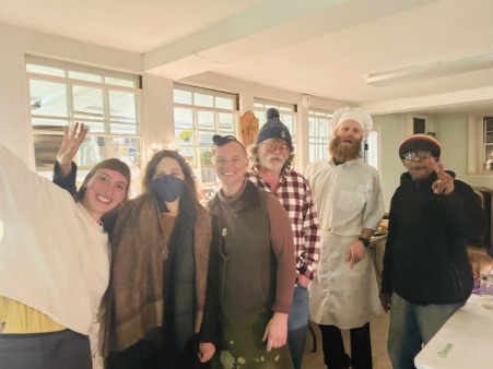 Primary and supportive chefs of weekly Plainfield Community Meal six individuals of different generations, race, and gender, standing next to each other and smiling in a softly lit church basement during Plainfield Community Meal.
