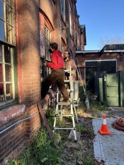 two people, one on a ladder and one standing to steady it, fixing the windows at arch st warehouse