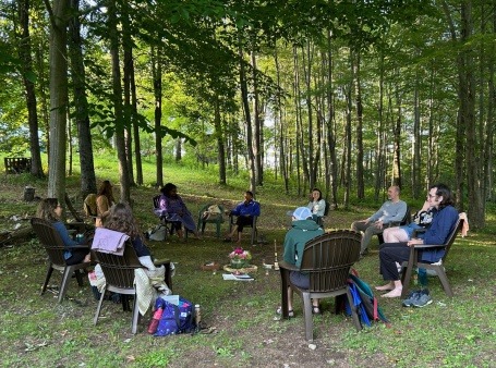 A diverse group of people gather in circle under a canopy of trees. 