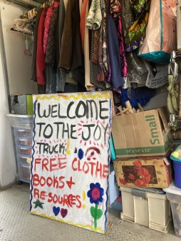 A large hand painted sign leans up against boxes of art supplies and the clothing rack inside the Joy Truck reading "Welcom to the Joy Truck Free Clothes, Resources and Art Supplies"