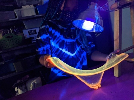 a black girl with braids and a tie dye shirt plays with orange slime that glows under the blacklight in front of her