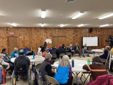 Residents gathered in a community center, listening to a panel of speakers at the front of the room.