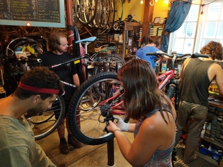 4 people, two at a workbench and two examining a bike nearby