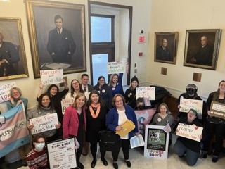 Over a dozen LGBTQ activists hold signs inside the state house. Behind them are portraits. Their signs say "live free means trans rights" "we support trans athletes" "ban guns not student athletes" "trans girls are girls" and "protect LGBTQ children now!" 