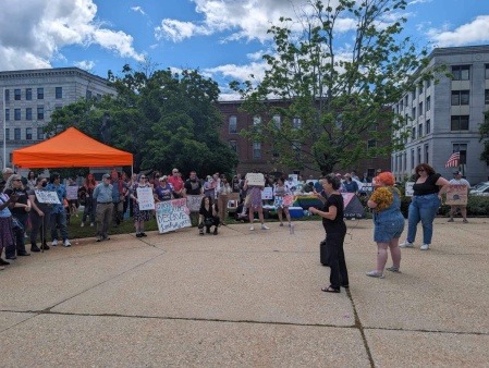 A crowd of LGBTQ activists forms a circle around an activist with bright orange hair speaking to the crowd. To the side of them is a sign language interpreter. 