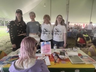 four members of 603 Equality stand behind a colorful table at Portsmouth Pride, with someone with pastel pink/purple hair knees at the table to write a letter